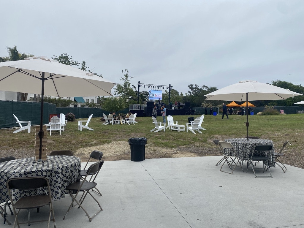 Outdoor event setup with chairs, umbrellas, stage, and scattered audience.