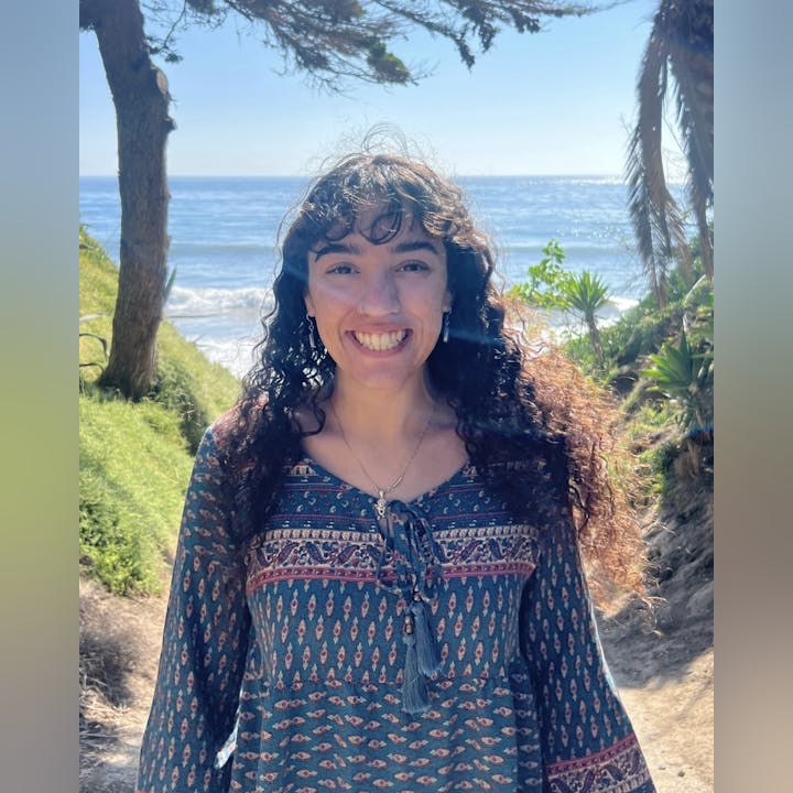 A smiling person with curly hair stands on a sunny beach path, surrounded by greenery and ocean views.