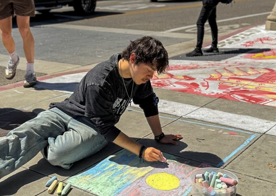 A person is creating sidewalk art with chalk, focusing on a colorful design featuring a sun. Other pedestrians are visible nearby.