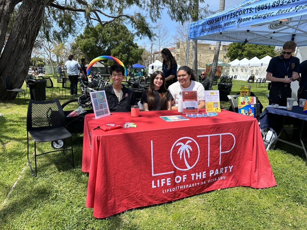 Three people behind a table with a "LIFE OF THE PARTY" banner at an outdoor event with tents and informational materials.