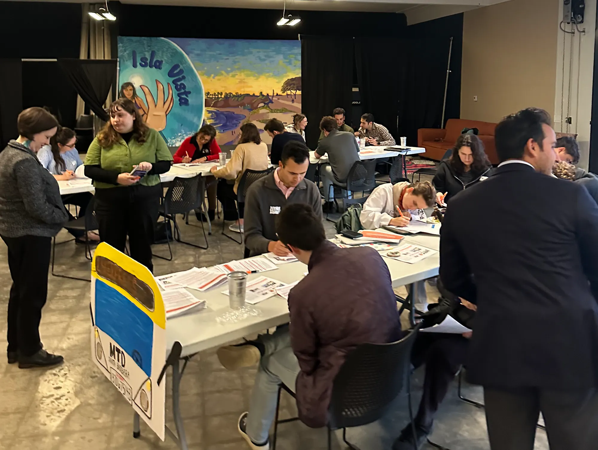 A busy indoor event with people seated at tables, some standing and conversing, and background artwork.