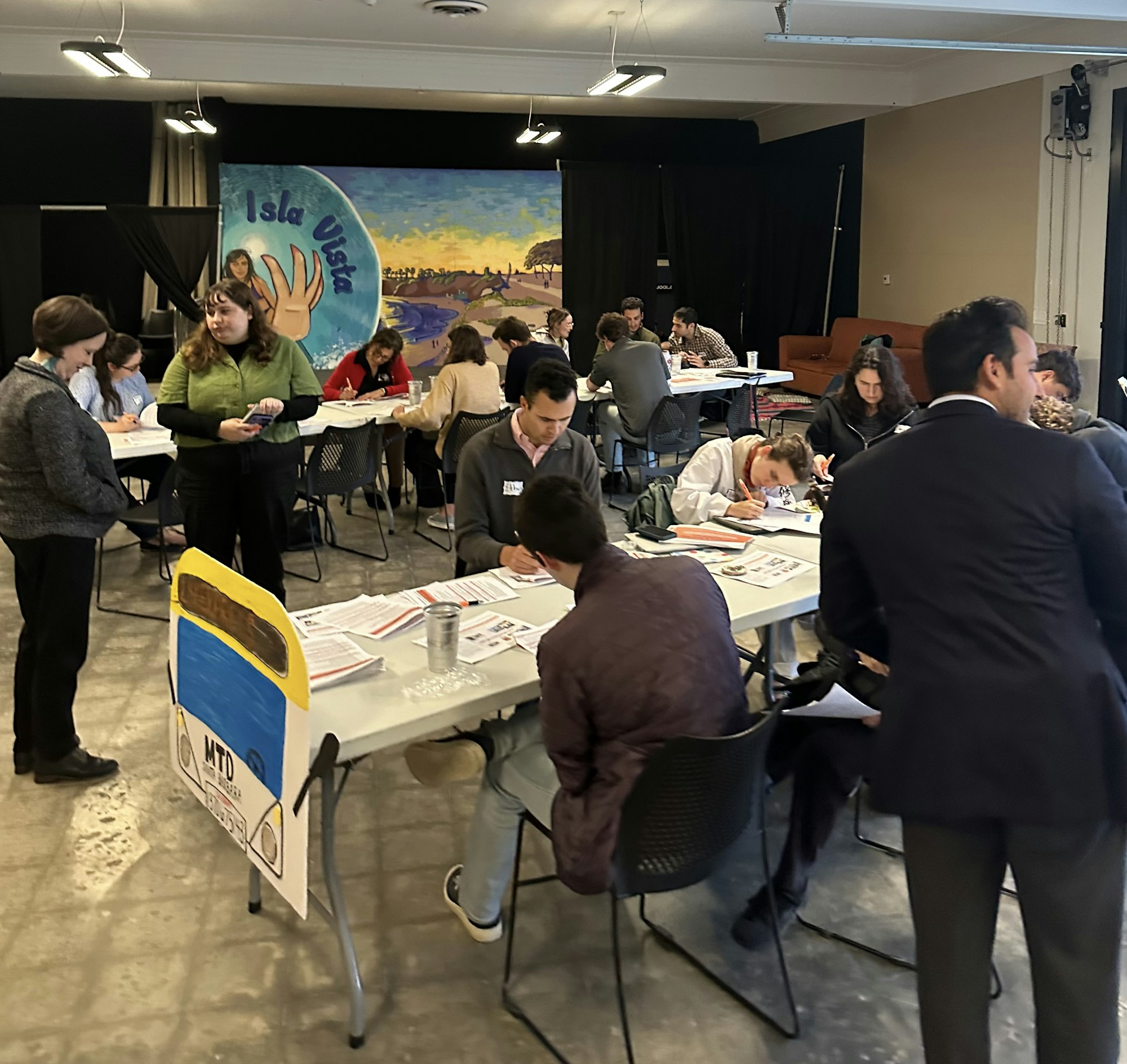 A busy indoor event with people seated at tables, some standing and conversing, and background artwork.