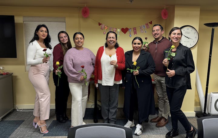 A group of seven people holding roses, smiling, with Valentine's Day decorations in the background.