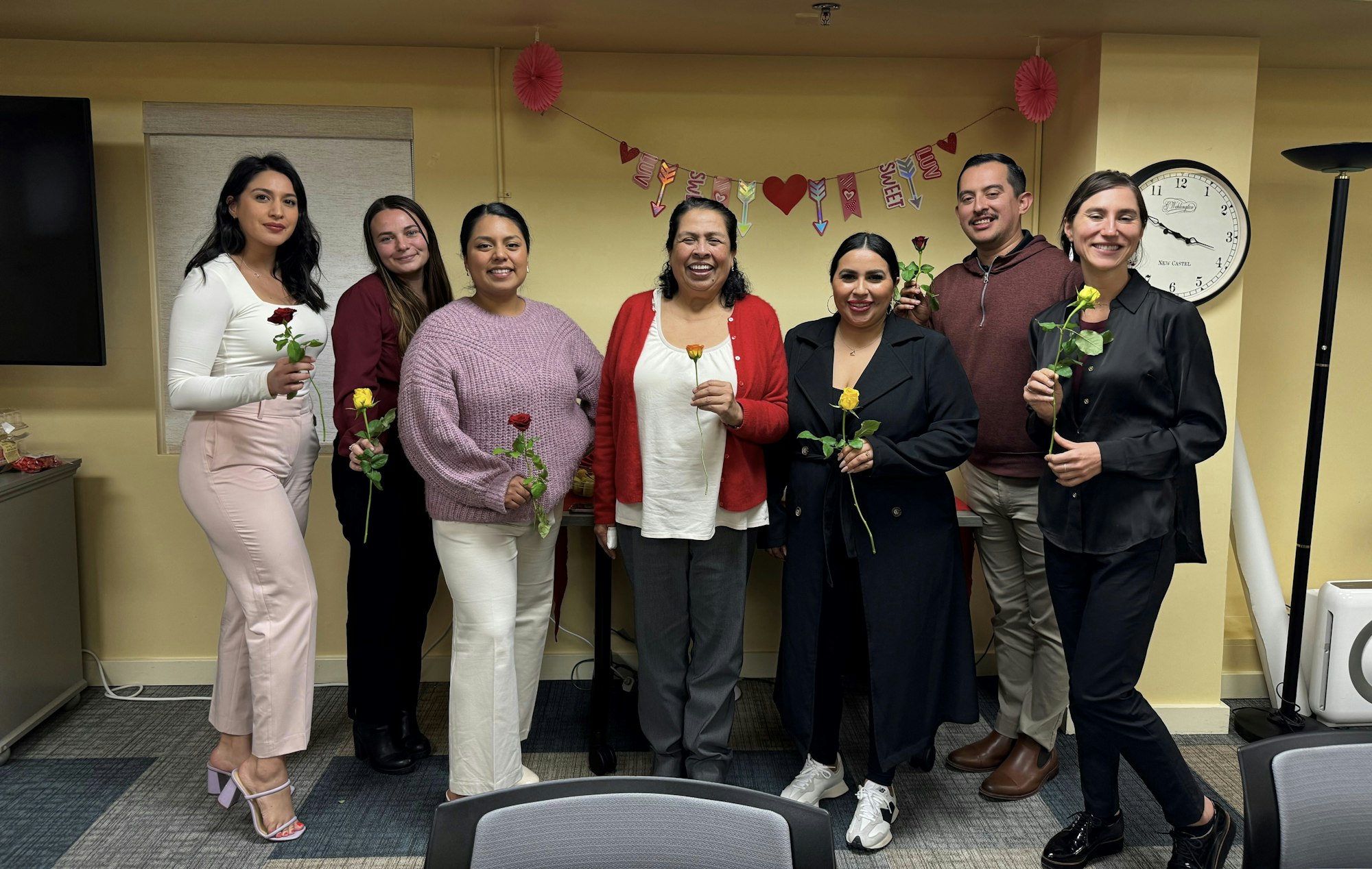 A group of seven people holding roses, smiling, with Valentine's Day decorations in the background.