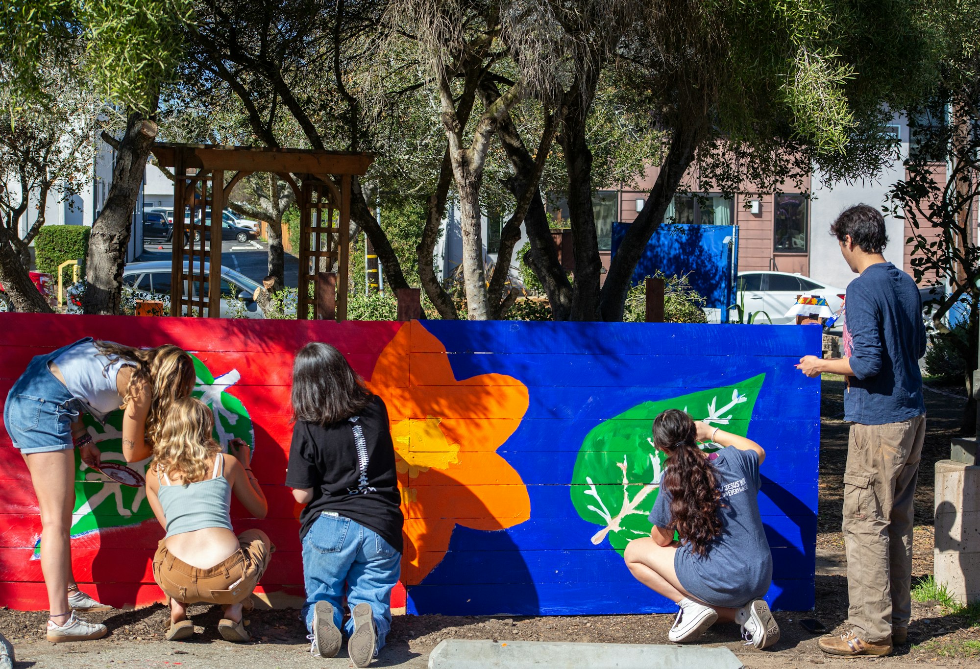 People painting a colorful mural with flowers and leaves on a wooden surface in a park setting.