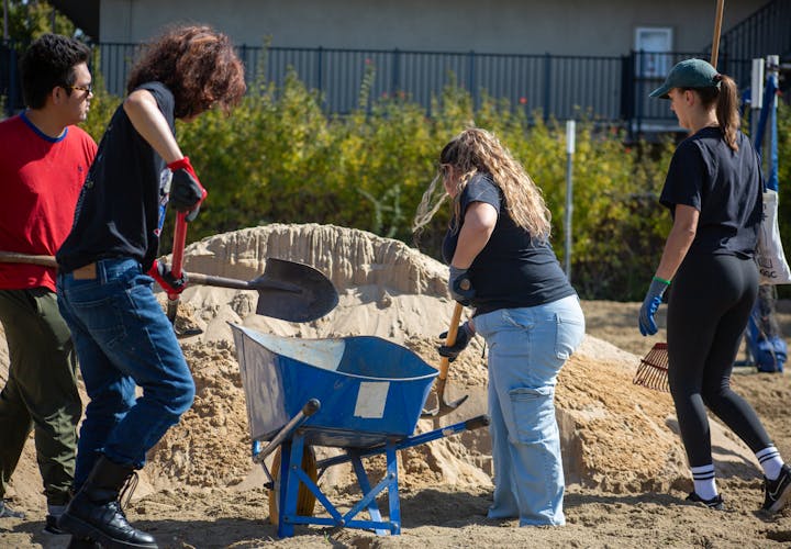 Four people using shovels and rakes, working with a blue wheelbarrow and sand.