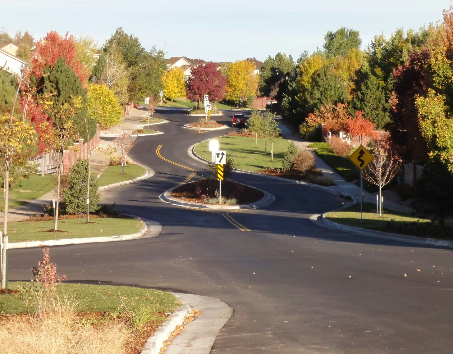 A winding road lined with autumn-colored trees and signs, set in a suburban neighborhood.