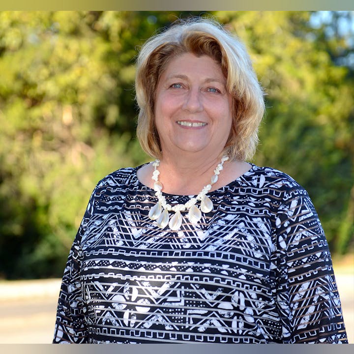 A smiling woman in a patterned top with a white necklace outdoors.