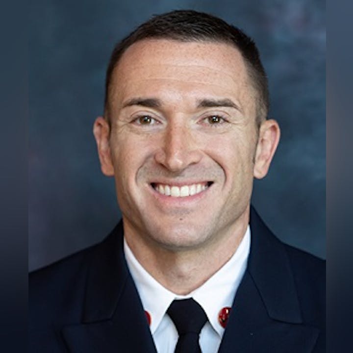 A smiling man in a dark suit with a lapel pin against a mottled blue background.