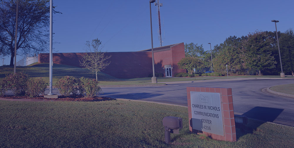 A building sign for "Charles W. Nichols Communications Center," lawn, shrubs, lamps, and a clear sky.