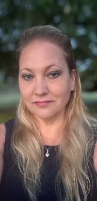 A woman with long blonde hair, wearing a black top, in front of a blurred natural background.