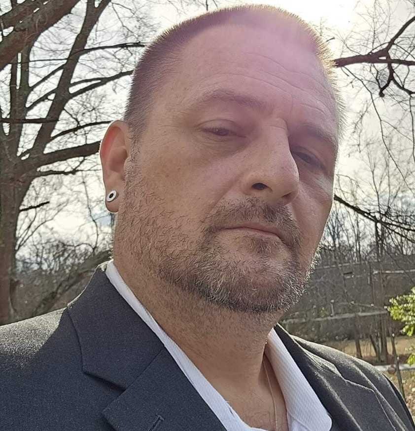 Man with a beard, ear gauge, and suit jacket outdoors.