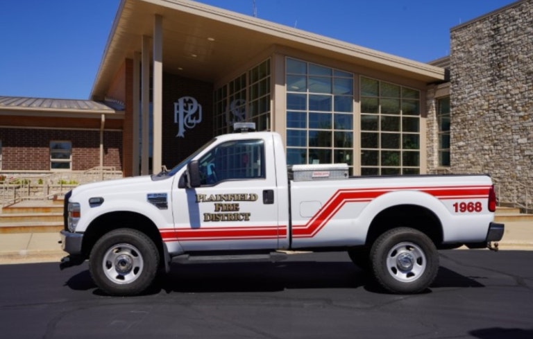 The image shows a white pickup truck marked as belonging to the Plainsfield Fire District, parked outside a building.