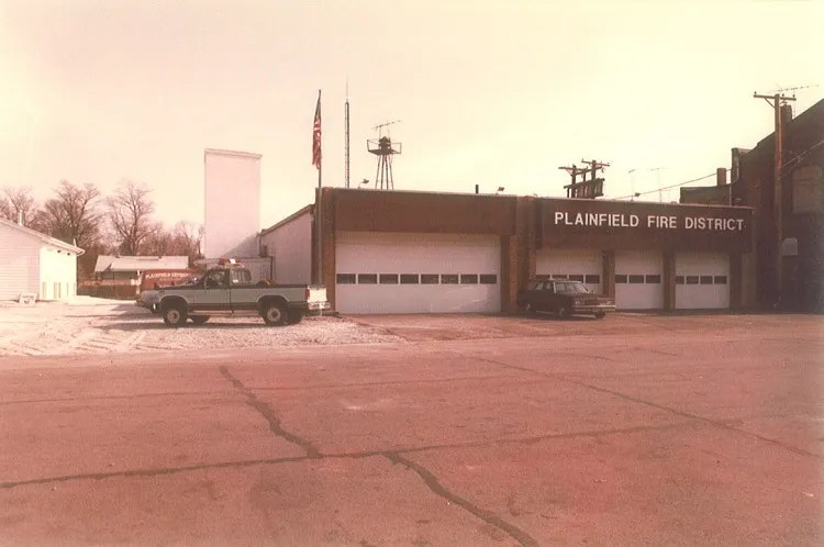 A fire station in Plainfield, featuring a truck and a car parked outside, surrounded by sparse trees and a quiet road.