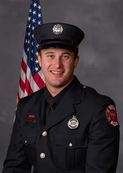 A smiling individual in a firefighter uniform stands in front of an American flag backdrop.