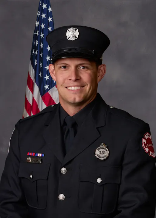 A smiling firefighter in uniform stands in front of an American flag, showcasing badges and decorations on his uniform.