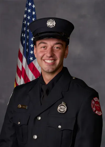 A smiling individual in a firefighter uniform stands in front of an American flag backdrop.