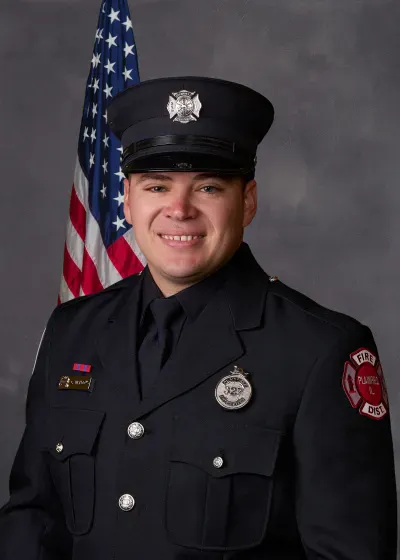 The image shows a firefighter in uniform, smiling, with an American flag background.