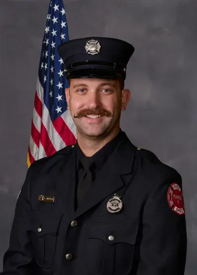 A smiling person in a firefighter uniform poses for a photo in front of an American flag.
