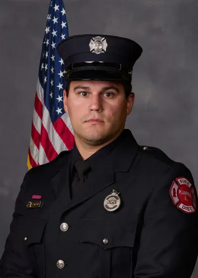 A male firefighter in uniform, posed in front of an American flag, with a serious expression.