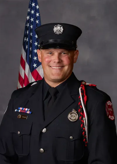 A firefighter in uniform smiling, standing in front of an American flag, highlighting dedication and service.