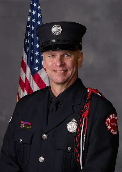 A person in a firefighter uniform is smiling against a background featuring an American flag.