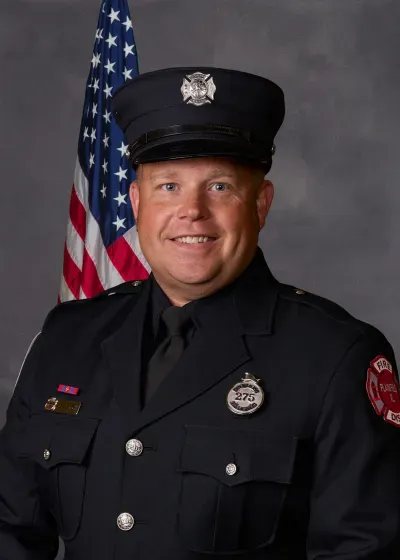 A smiling individual in a firefighter uniform, standing in front of an American flag backdrop.