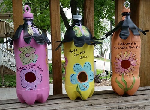 Three painted plastic bottles repurposed as decorative birdhouses, adorned with flowers and cheerful phrases.