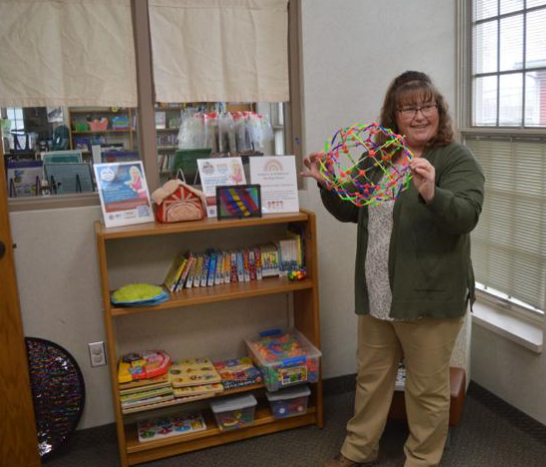 A woman holding a colorful expandable ball stands beside a shelf with books and educational toys.