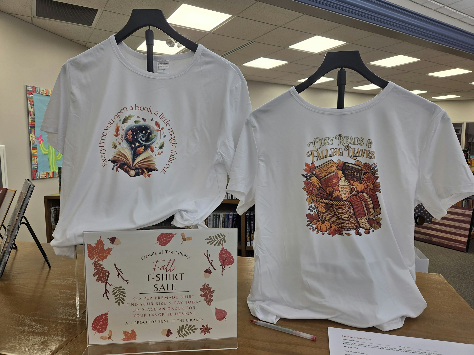 The image shows two fall-themed T-shirts for sale, featuring book-related designs and a sign promoting a library fundraiser.