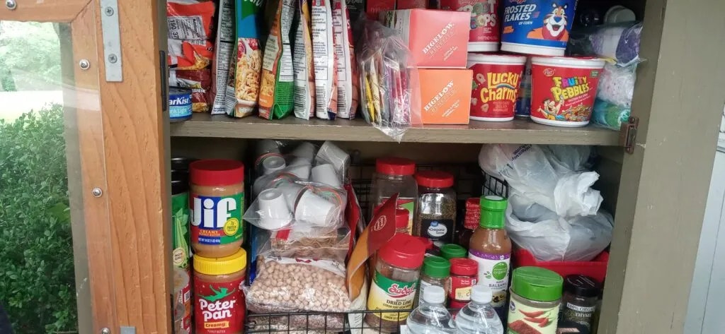 The image shows a pantry shelf crowded with various snacks, cereals, spices, and jars, organized within a wooden cabinet.