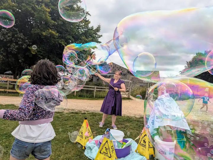 A woman creates large bubbles while a child watches, surrounded by bubble-making supplies and caution signs in a park.