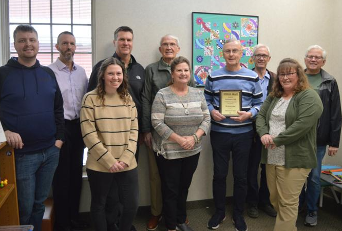 A group of people, some smiling, posing in a room. One person holds a plaque. There's colorful artwork on the wall behind them.
