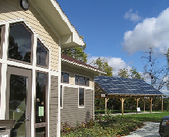 A building with large windows and a solar panel array on a stand outside, set against a backdrop of a clear blue sky with clouds.