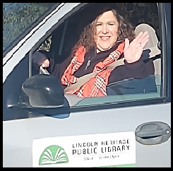 A person waving from a vehicle with a "Lincoln Heritage Public Library" sign on the door.