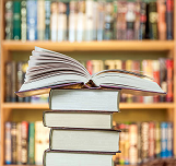 A stack of books with one open on top, set against a blurred bookshelf background.