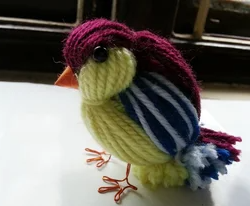 A colorful yarn bird with a purple head, striped wings, and wire legs.