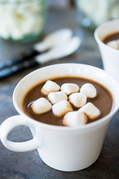 A cup of hot chocolate topped with marshmallows, set against a blurred background featuring more marshmallows and utensils.
