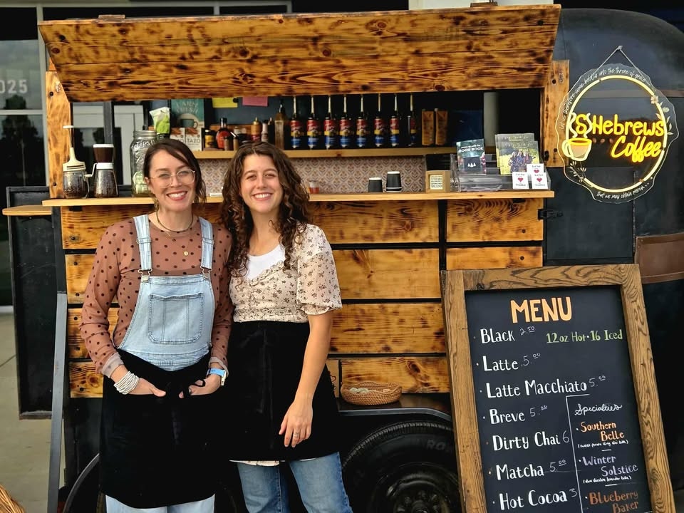 Two women stand in front of a coffee stand named "Hebrews Coffee," showcasing a menu with various coffee options.