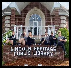 Six people sitting on a sign reading "Lincoln Heritage Public Library" in front of a brick building.
