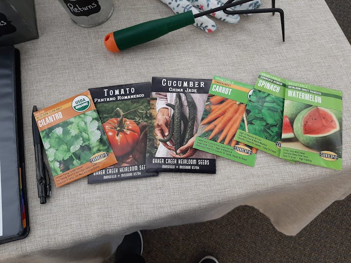 Packets of seeds for cilantro, tomato, cucumber, carrot, spinach, and watermelon on a table with a garden tool.
