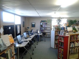 A library with bookshelves, computers, and plants under fluorescent lighting.