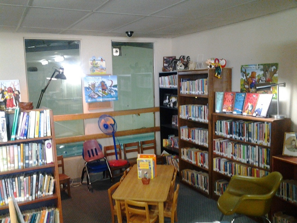 A children's library section with bookshelves, colorful posters, a table with a chair, and a reading lamp.
