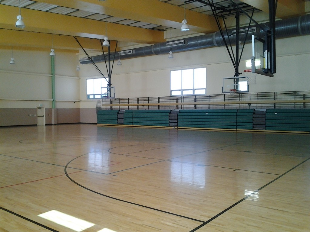An empty indoor basketball court with a hoop, bleachers, and high ceiling.