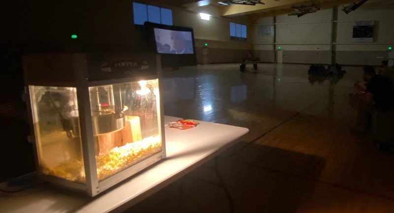 Popcorn machine on a table with a movie being projected in a dimly lit gymnasium, people sitting on the floor watching.