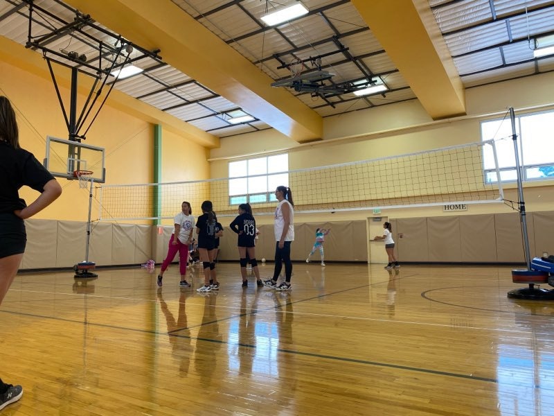 People playing volleyball indoors, with a basketball hoop in the background.