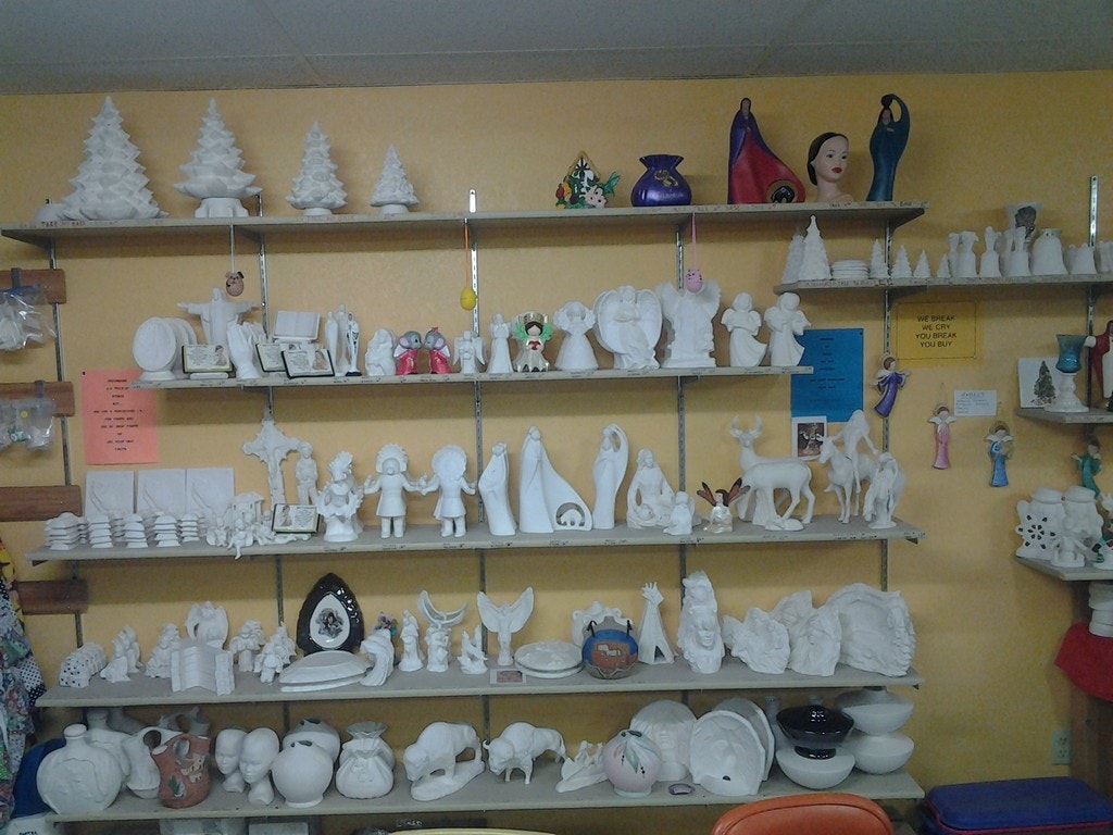 Shelves with unpainted ceramics, figurines, and decor items in a craft shop with a "you break, you buy" sign.