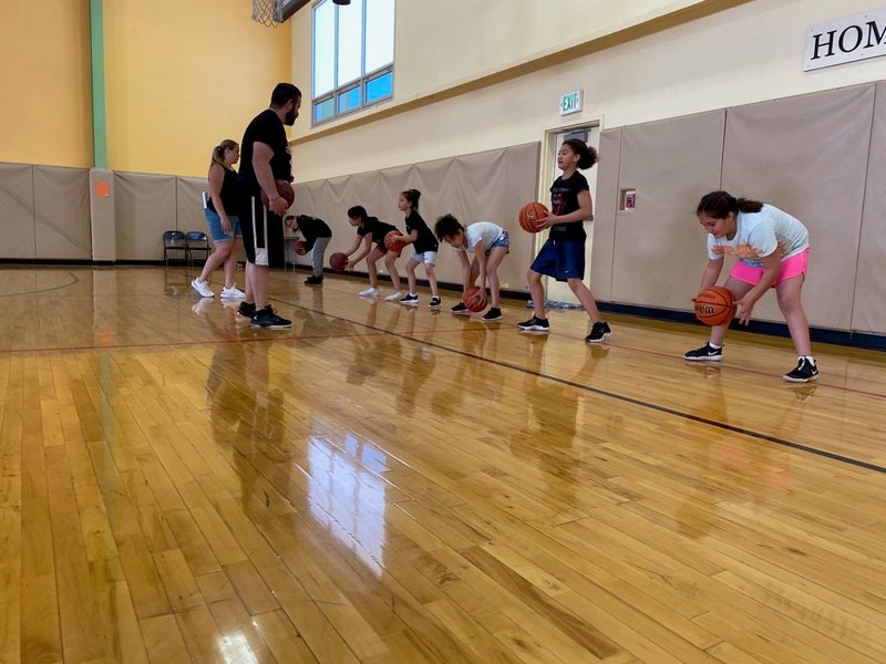 Basketball practice, kids dribbling balls in a gym, with a coach supervising.