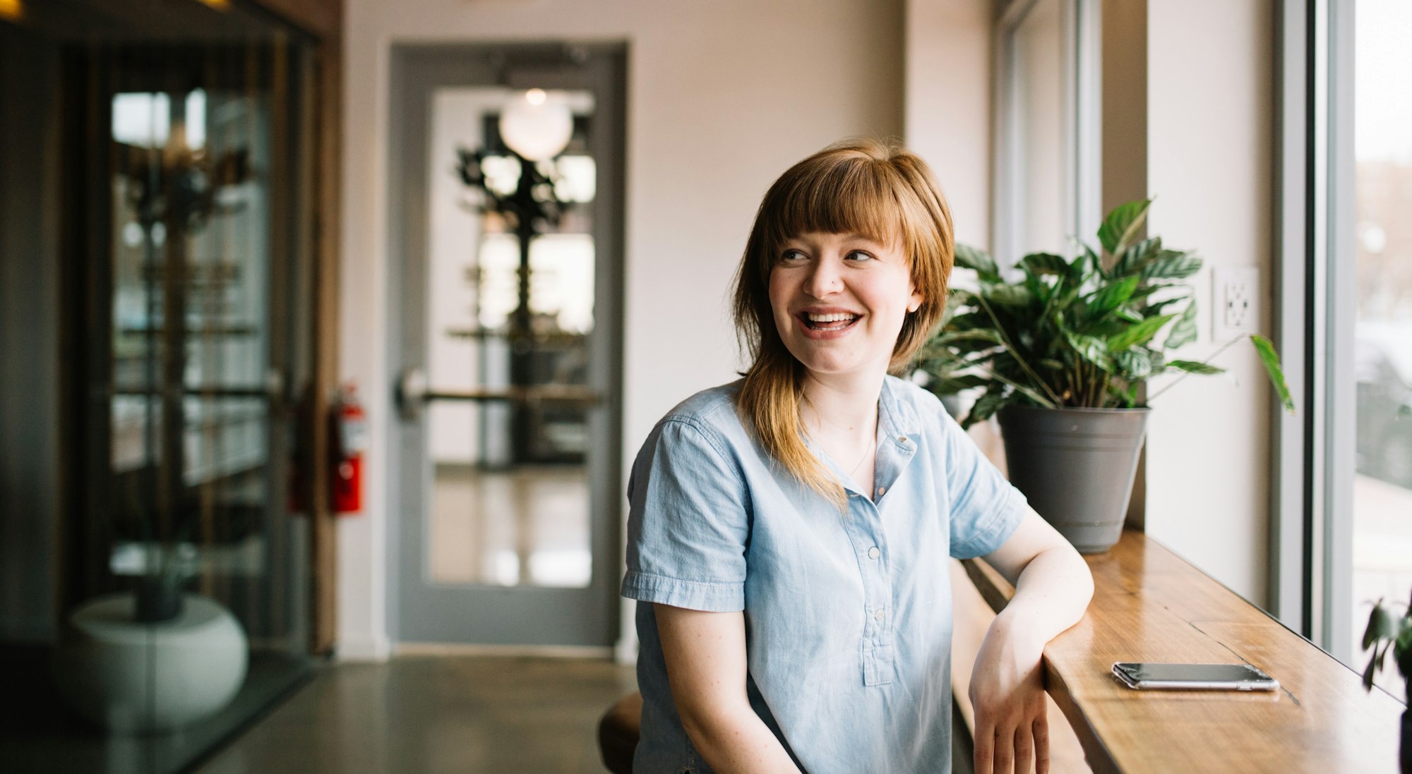 A smiling woman in a casual blue shirt sits by a window, with plants and a phone on the wooden counter beside her.