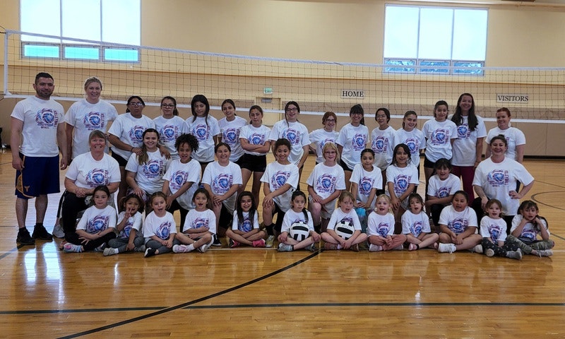 Group of people in matching shirts posing in a gym with a volleyball net.